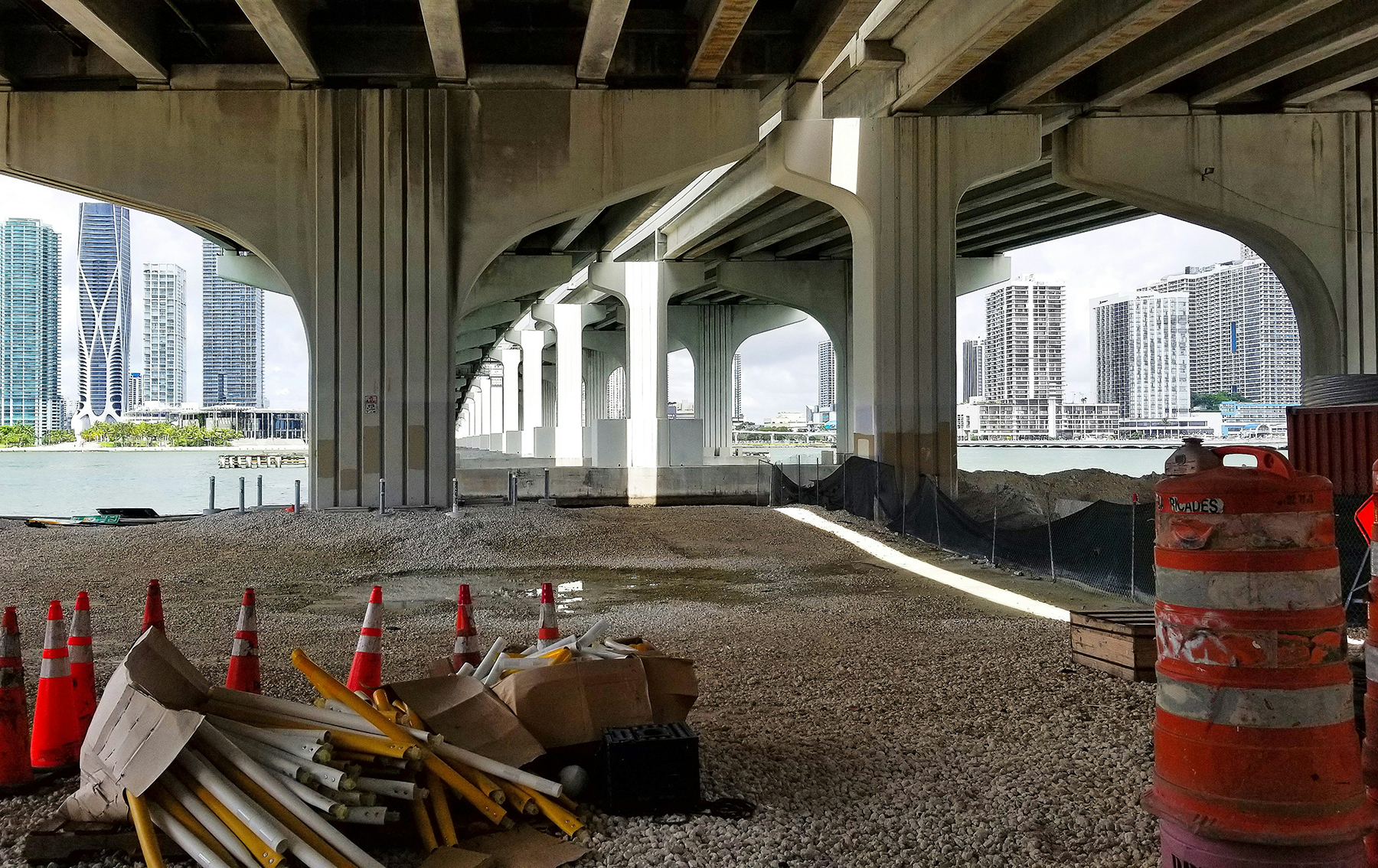 image of the underside of a bridge with traffic cones and pylons in the foreground and a water body and skyscrapers in the background image of the underside of a bridge with traffic cones and pylons in the foreground and a water body and skyscrapers in the background
