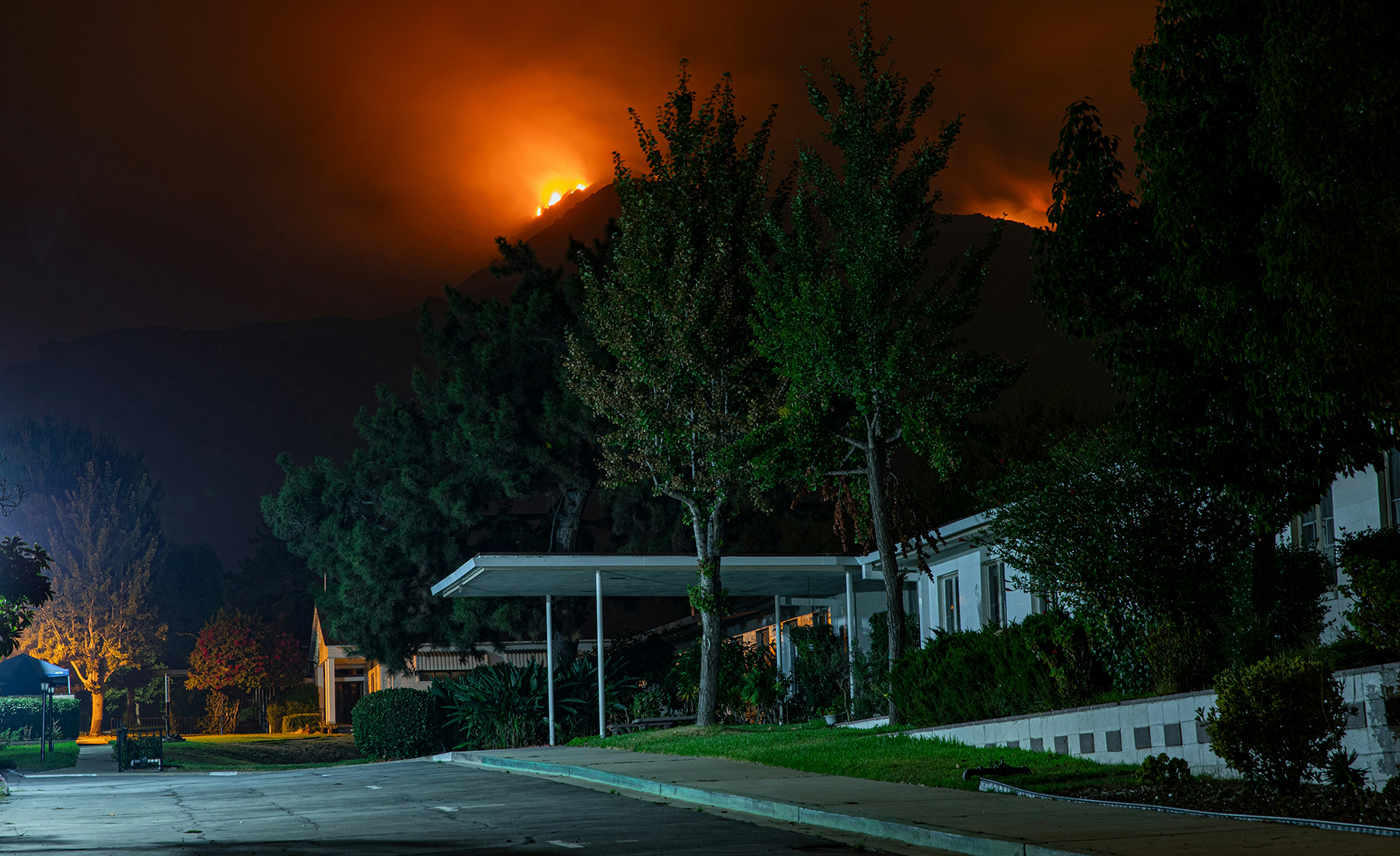 a house with green trees sits in the foreground while a wildfire rages in the distance in the background