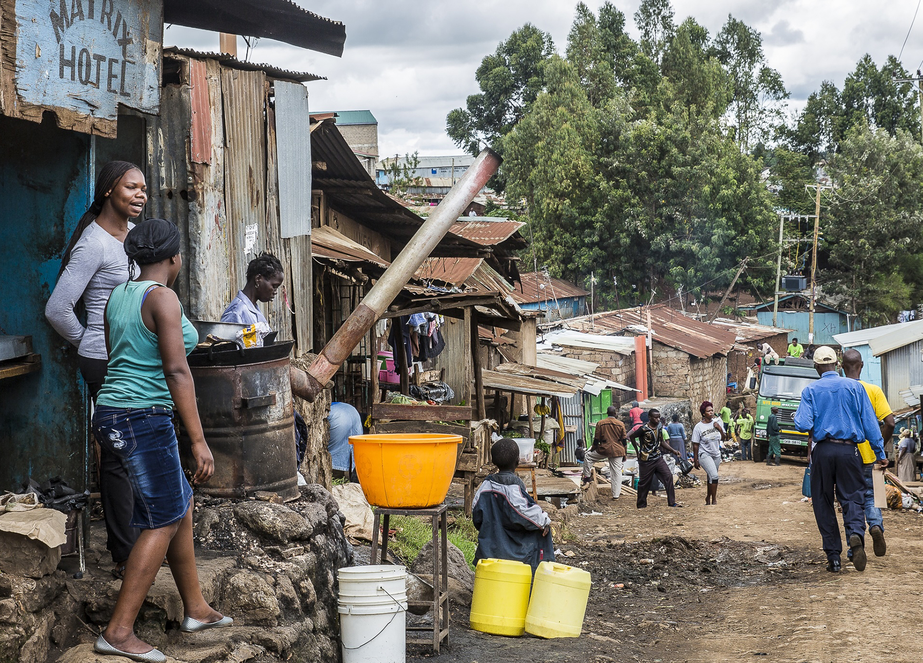 people walk down a dirt road with others standing near dilapidated buildings