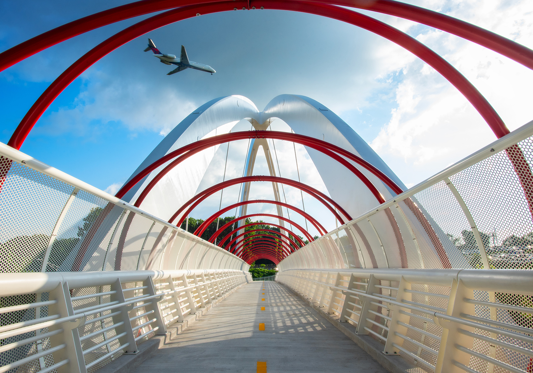 an airplane flies above a red and white pedestrian bridge