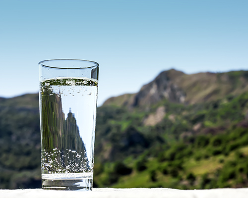 A clear glass of water with blue sky and green mountain landscape background 