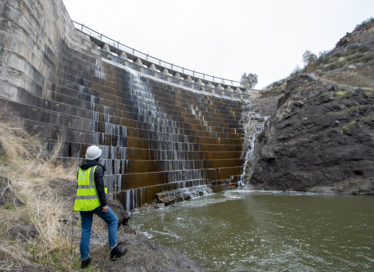 a person in a green safety vest and a hardhat observes water flowing from a dam