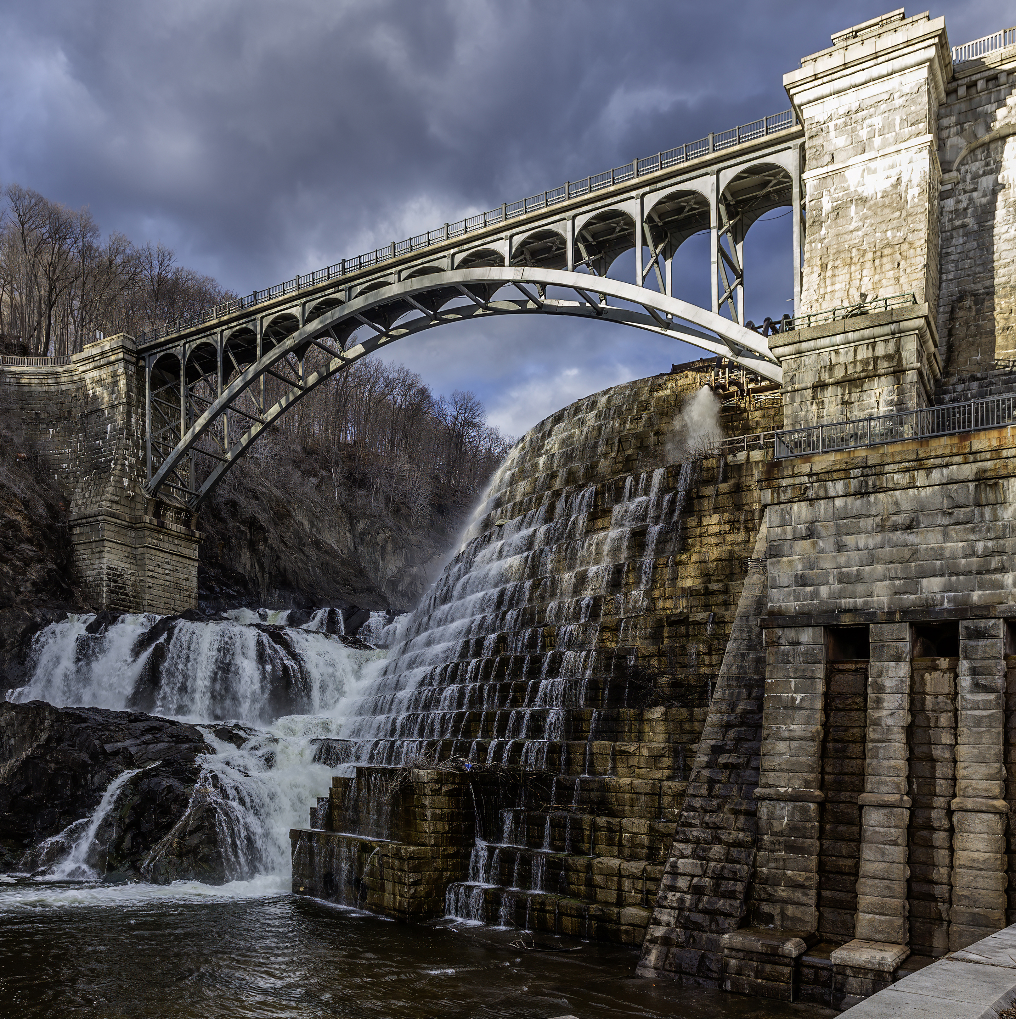 Water cascades over stone dam beneath arched bridge and cloudy sky