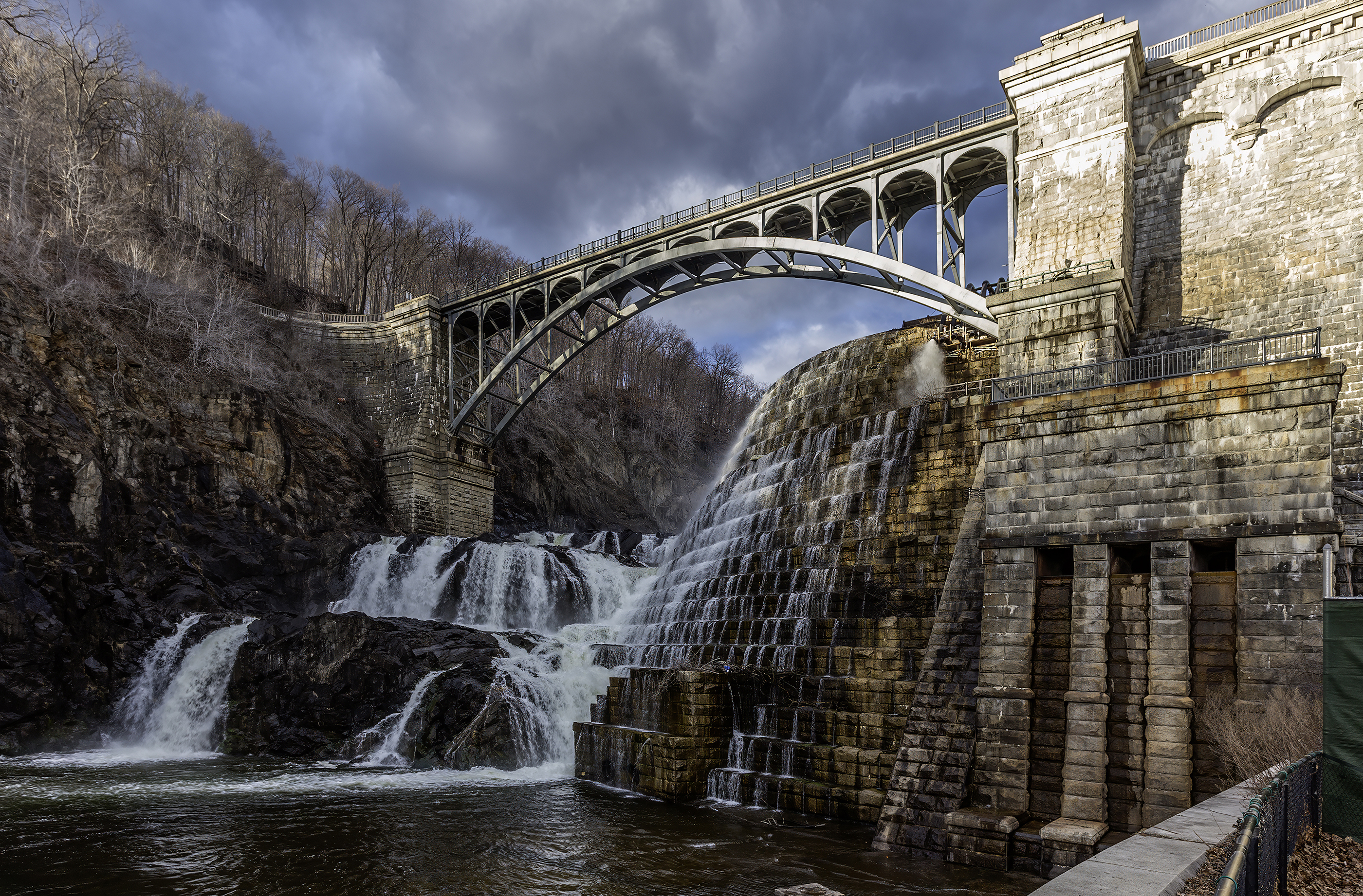 Water cascades over stone dam beneath arched bridge and cloudy sky
