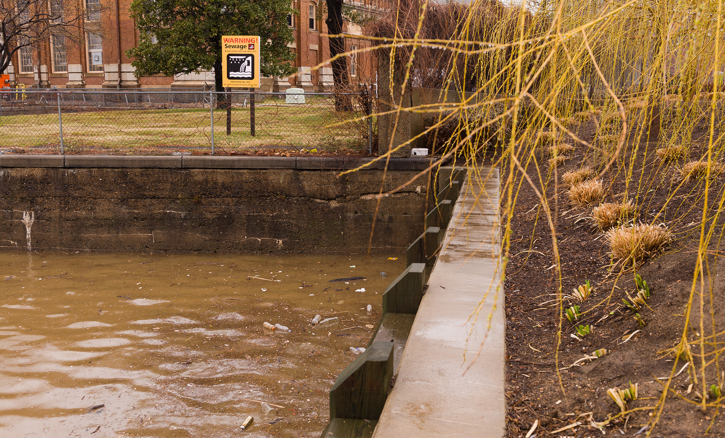 Sewage warning sign by polluted urban canal with debris in water