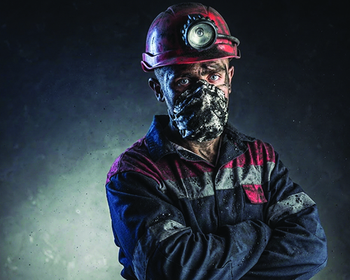 Man covered in coal dust with arms crossed wearing red hardhat, face mask, and work suit