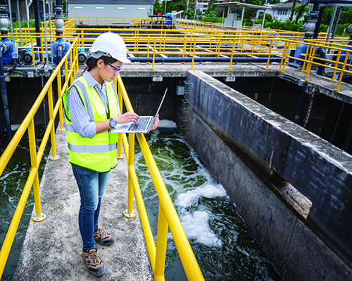 At a water treatment facility, a worker in a yellow safety vest and hardhat looks at a laptop