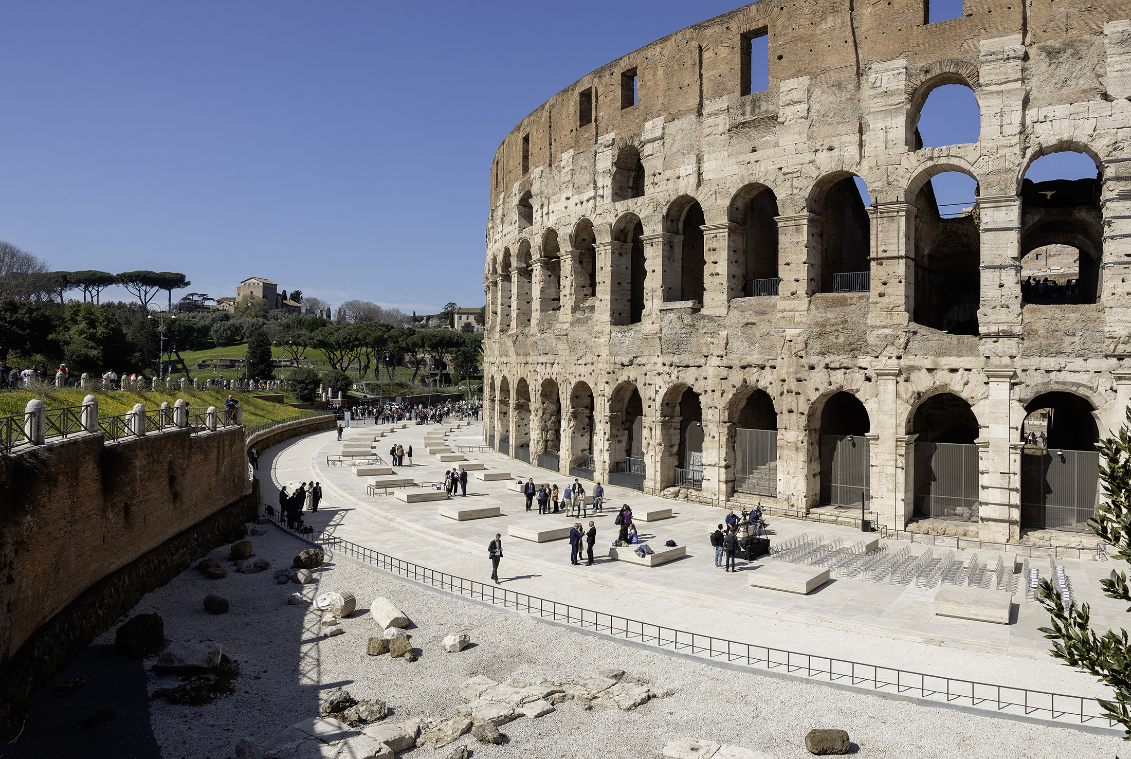 Colosseum exterior with restored plaza, visitors, and seating blocks underneath arches