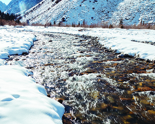 A clear mountain stream winds through a snow-covered valley on a bright day.