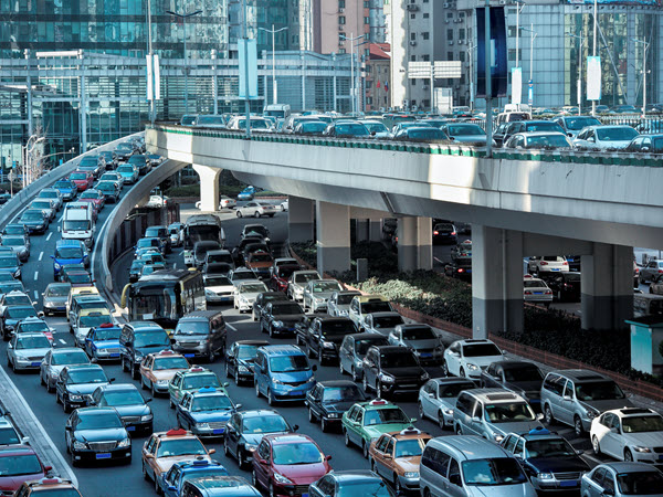 A dense traffic jam clogs a wide city street under a concrete overpass with city office buildings in the background